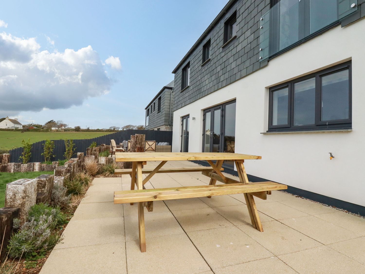 A patio with a wooden picnic table next to a modern building with large windows and a garden at Lavender House in Crantock