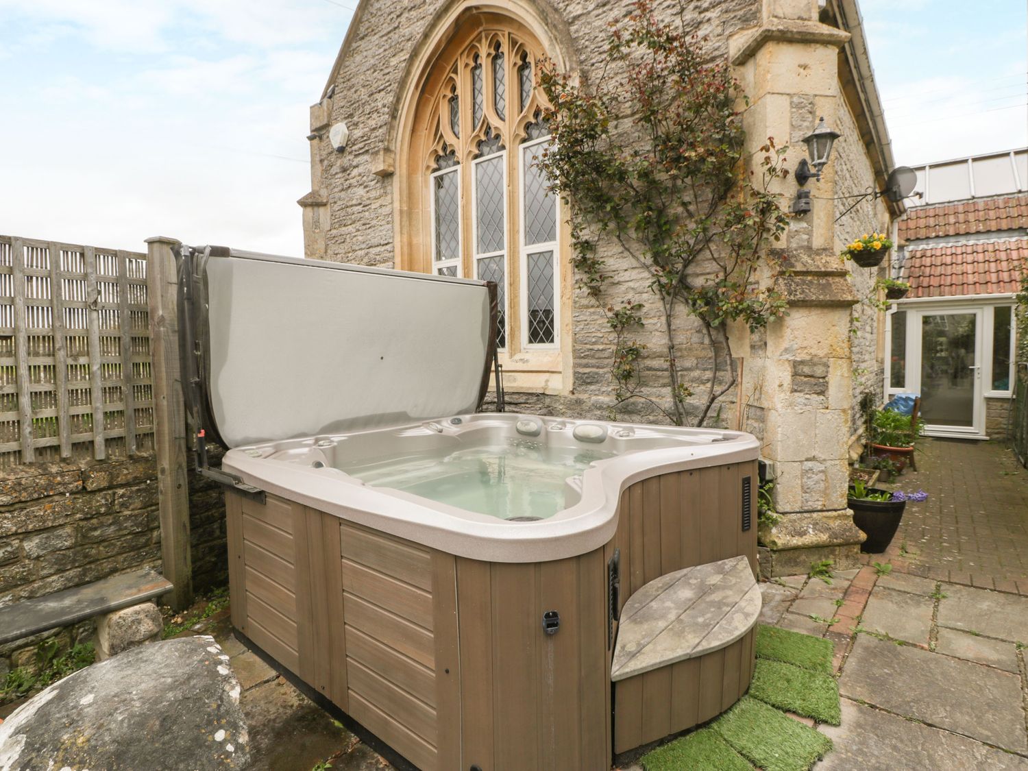 An outdoor hot tub next to a stone building with arched windows and a small garden path at The Coach House in Charlton Adam
