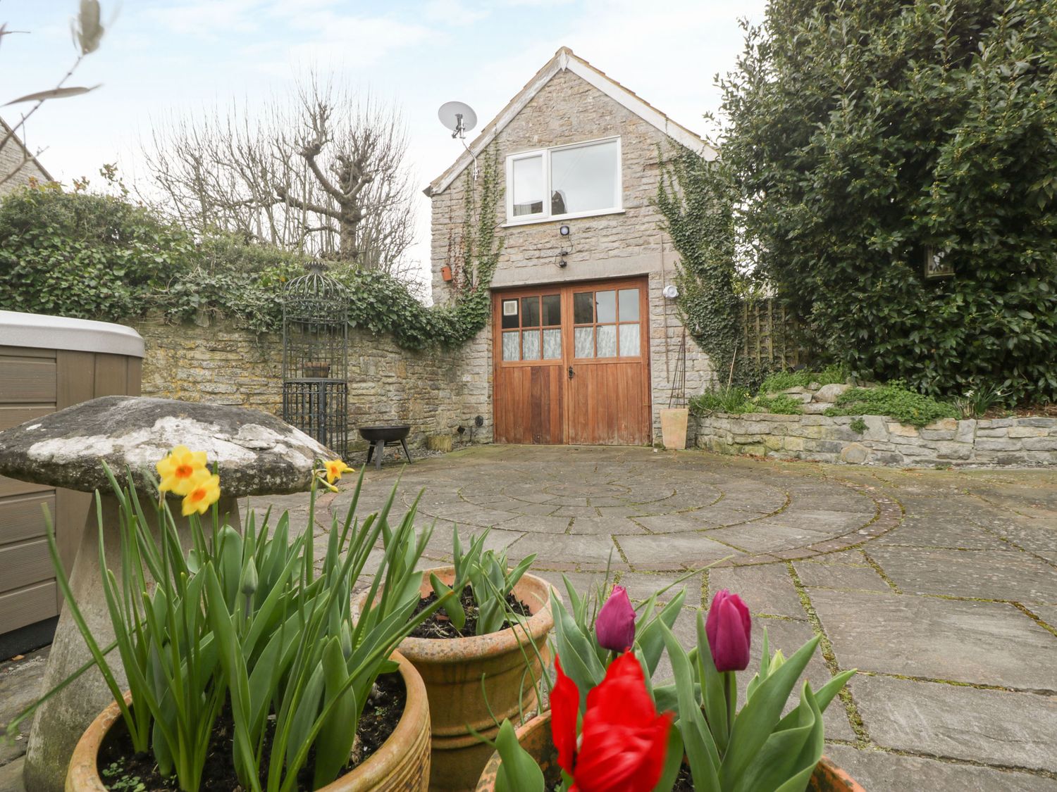 An outdoor area with potted flowers a stone patio and a stone building with wooden doors and a window at The Coach House in Charlton Adam