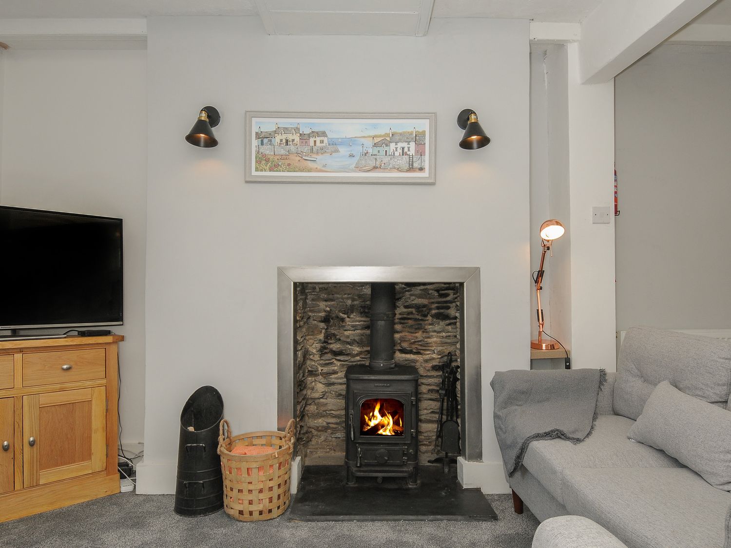 A living room with a fireplace a wood stove a gray sofa a television on a wooden cabinet and a lamp at Farleigh Cottage in Looe