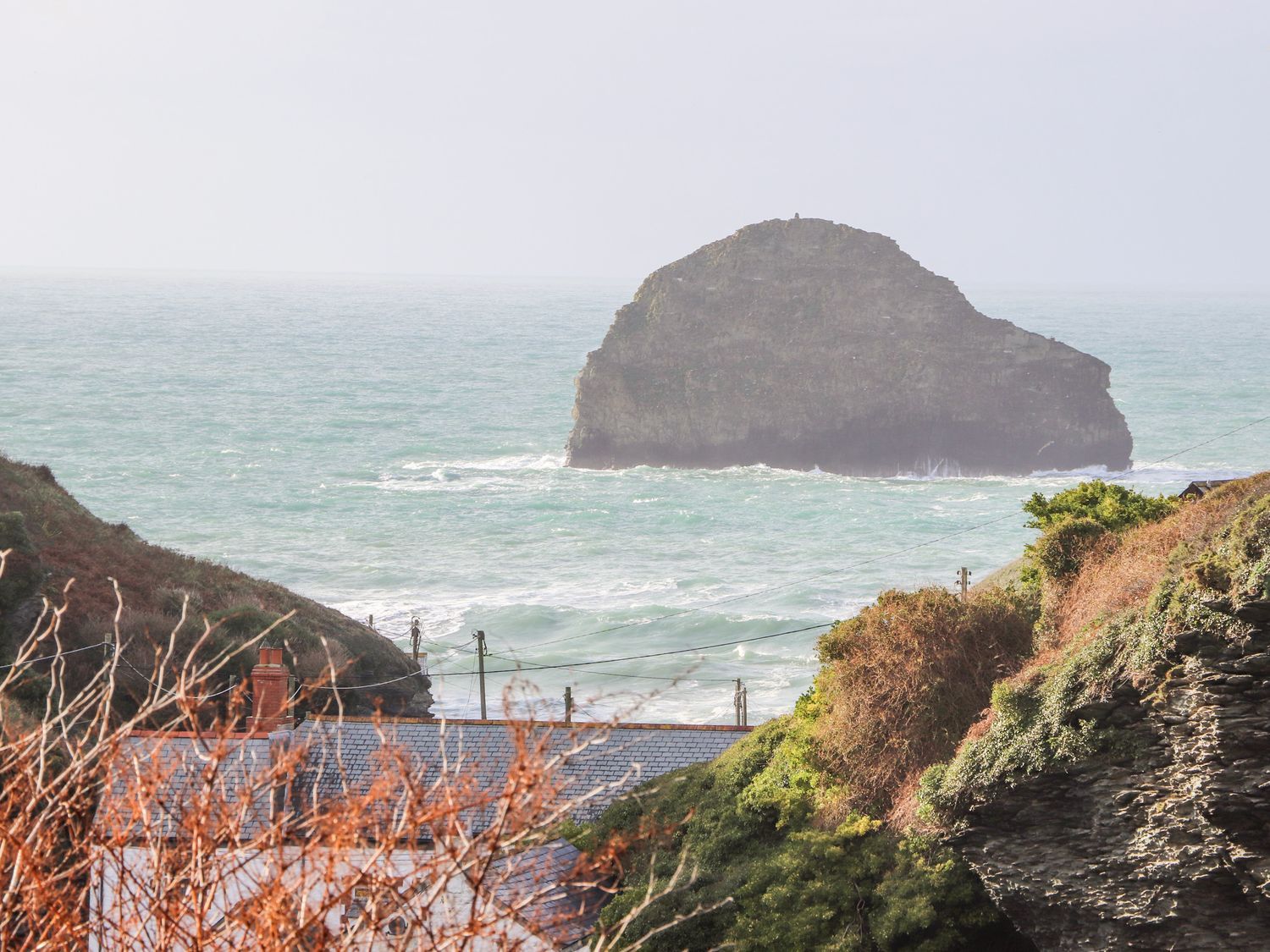 A coastal scene with a large rock formation in the sea and rooftops and vegetation in the foreground at Cliffside in Trebarwith Strand near Tintagel
