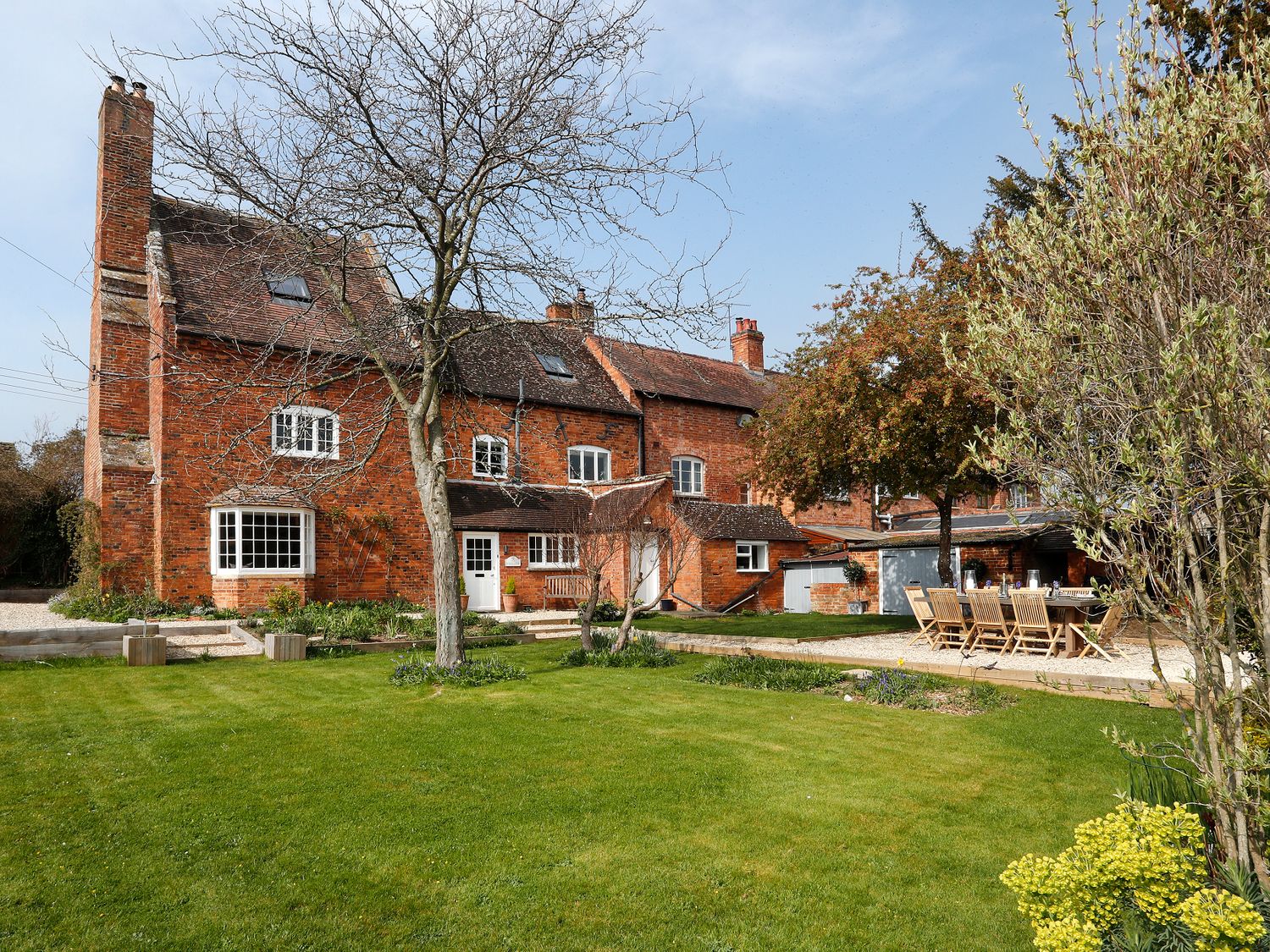 A large brick house with multiple chimneys a tree in front and an outdoor dining area with wooden furniture on a lawn at The Old School House in Dumbleton