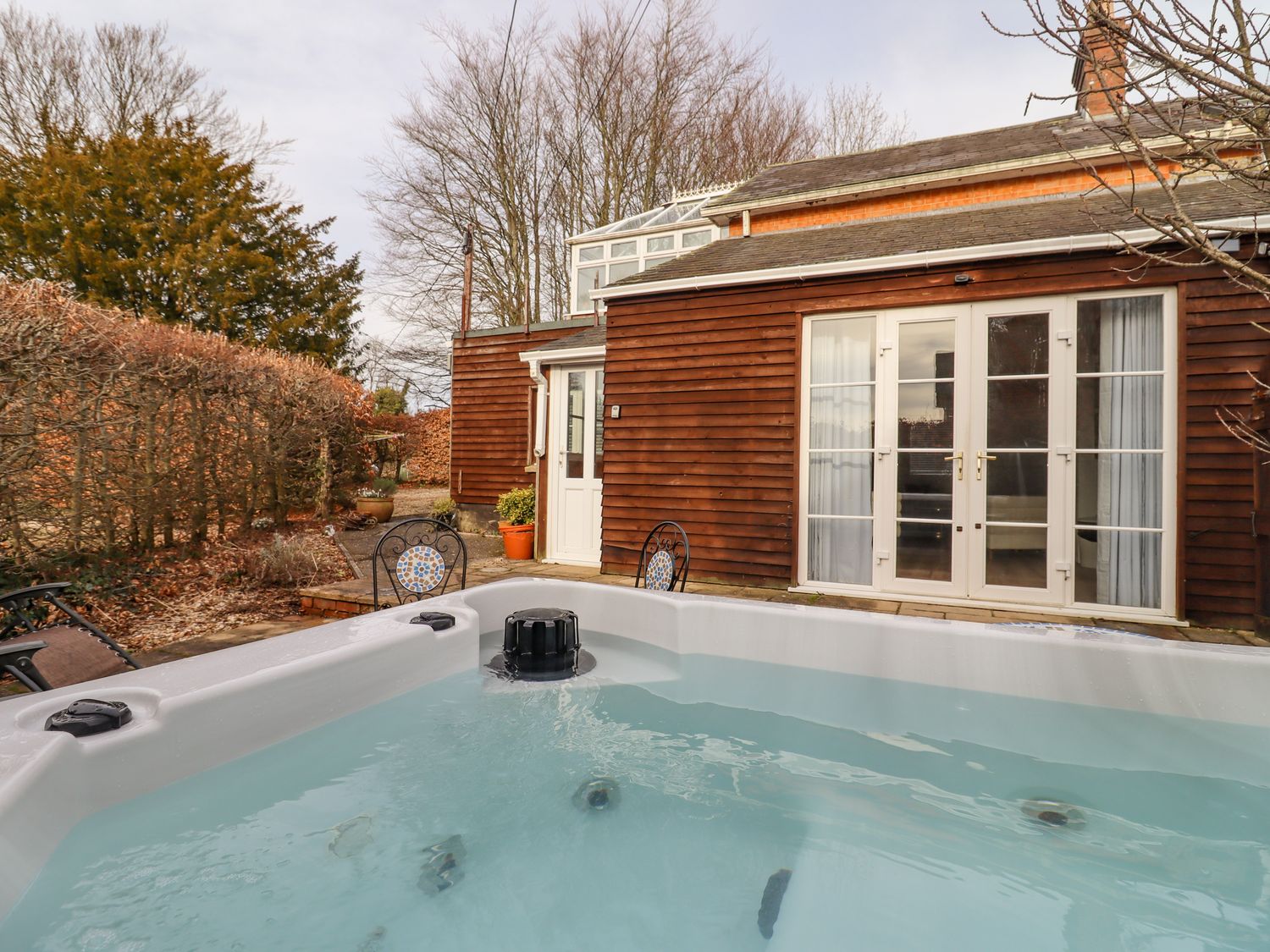 An outdoor hot tub with water in a backyard next to a wooden house with glass double doors and chairs