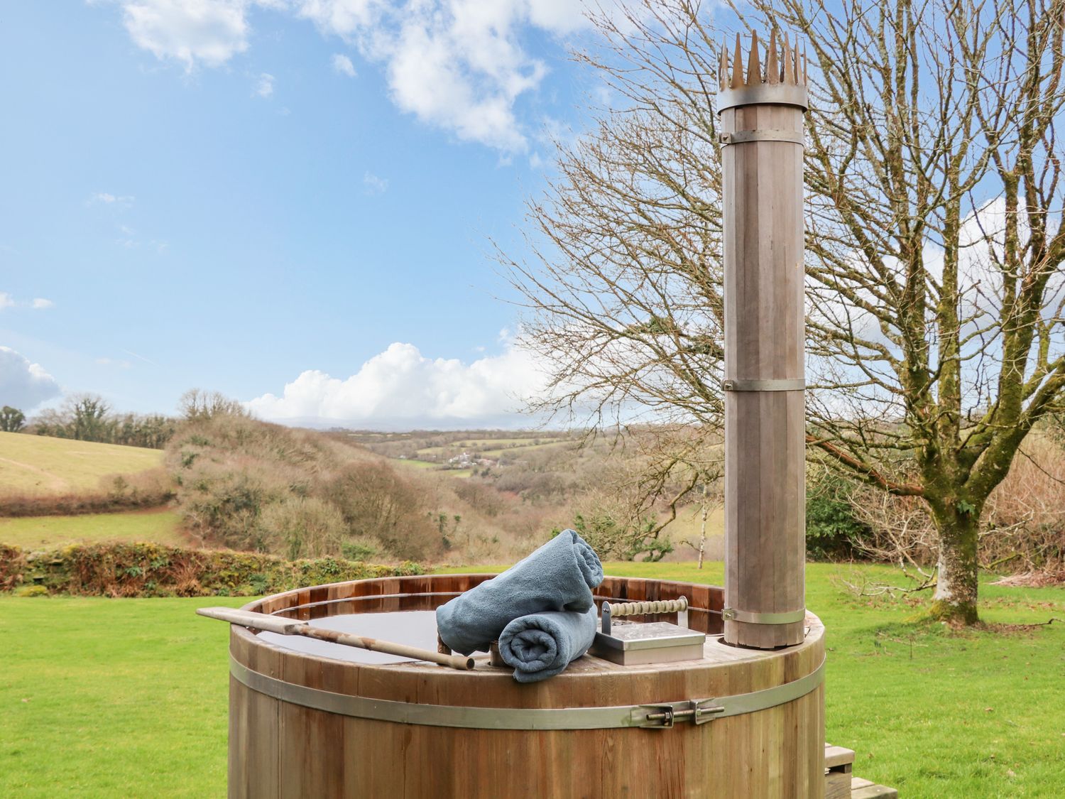 A wooden hot tub with towels on it outside on grass with trees and hills in the background at Treveddoe Farmhouse in Bodmin Moor