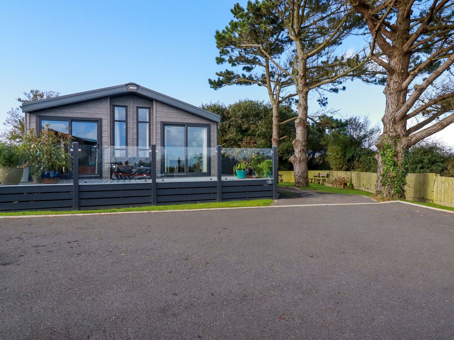 A grey cabin with large windows and a glass deck railing beside trees and a paved area at Piran Lodge in Padstow