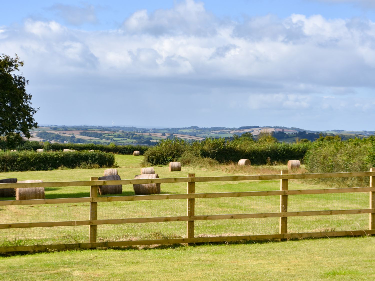 A field with hay bales and a wooden fence with hills and trees in the background at The Linhay in Lapford