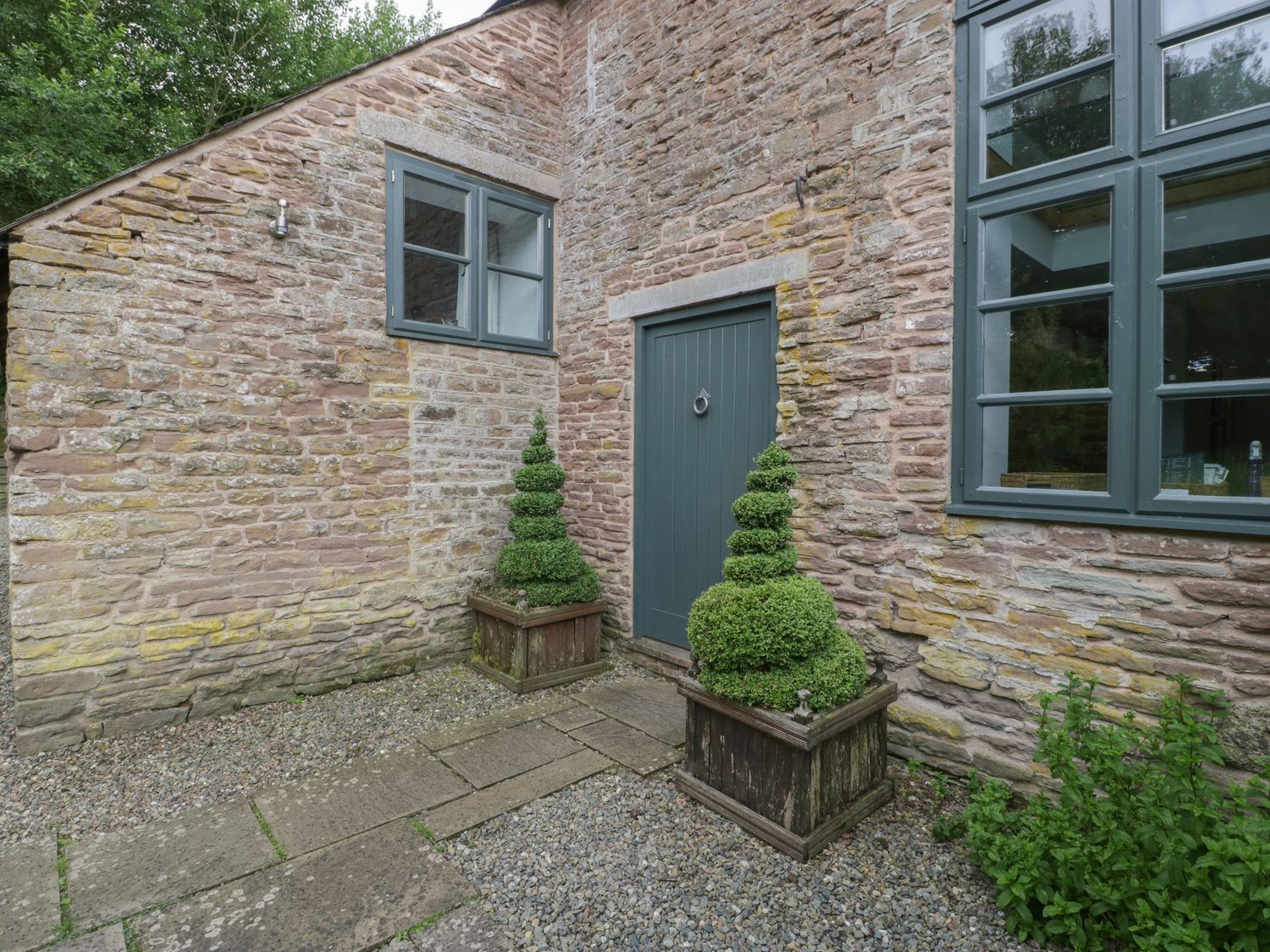 A stone building exterior with two windows a green door and two spiral-shaped topiary plants in wooden planters at Owl Barn Marston Stannett near Leominster