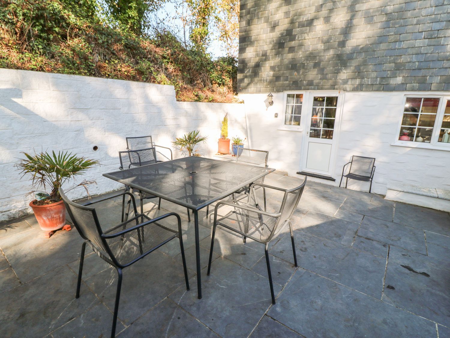 An outdoor patio area with a metal table and six chairs near a white wall and door at Beach Way Cottage in Gorran Haven