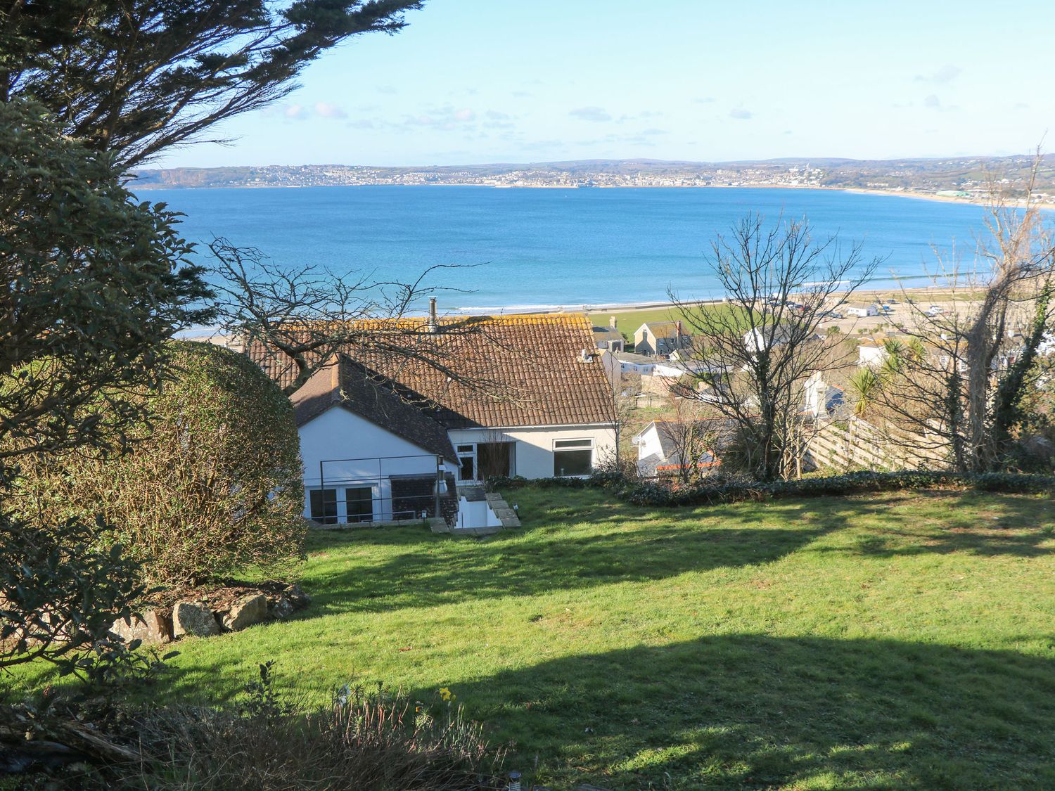 A garden with grass trees and bushes overlooking houses and a bay with water and hills in the background at Rosemount in Marazion