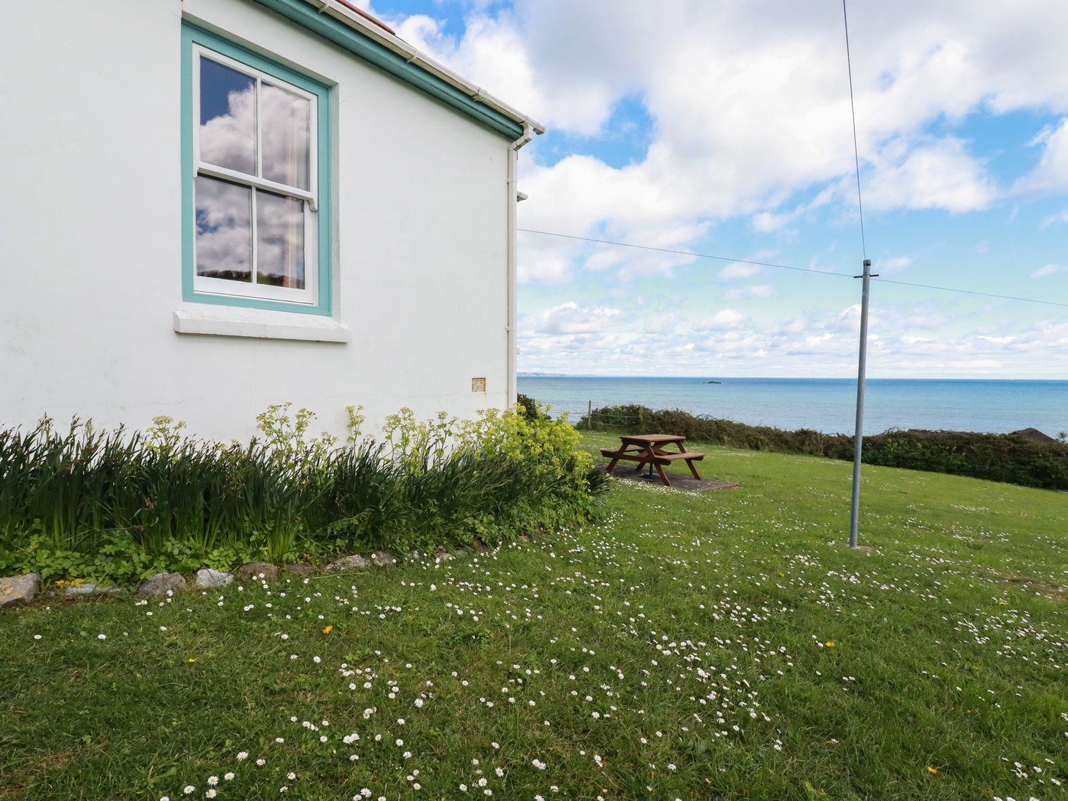 Side of a white house with a window and garden next to a lawn with a picnic table and a sea view at Beachcliff in Gorran Haven