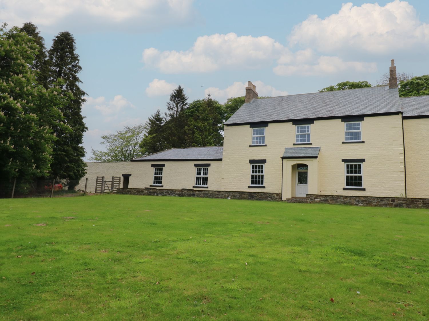 A two-story cream house with multiple windows and a green lawn surrounded by trees at Nenthead House in Nenthead near Alston