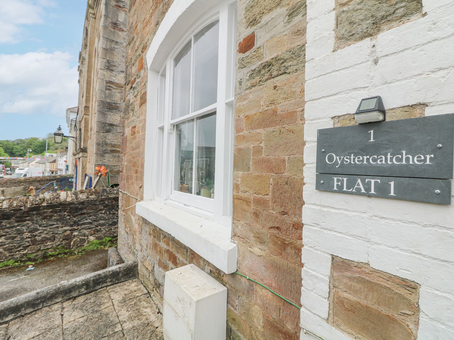 Exterior view of a brick building with a white framed window and a sign that reads Oystercatcher Flat 1 at The Oystercatcher in Falmouth