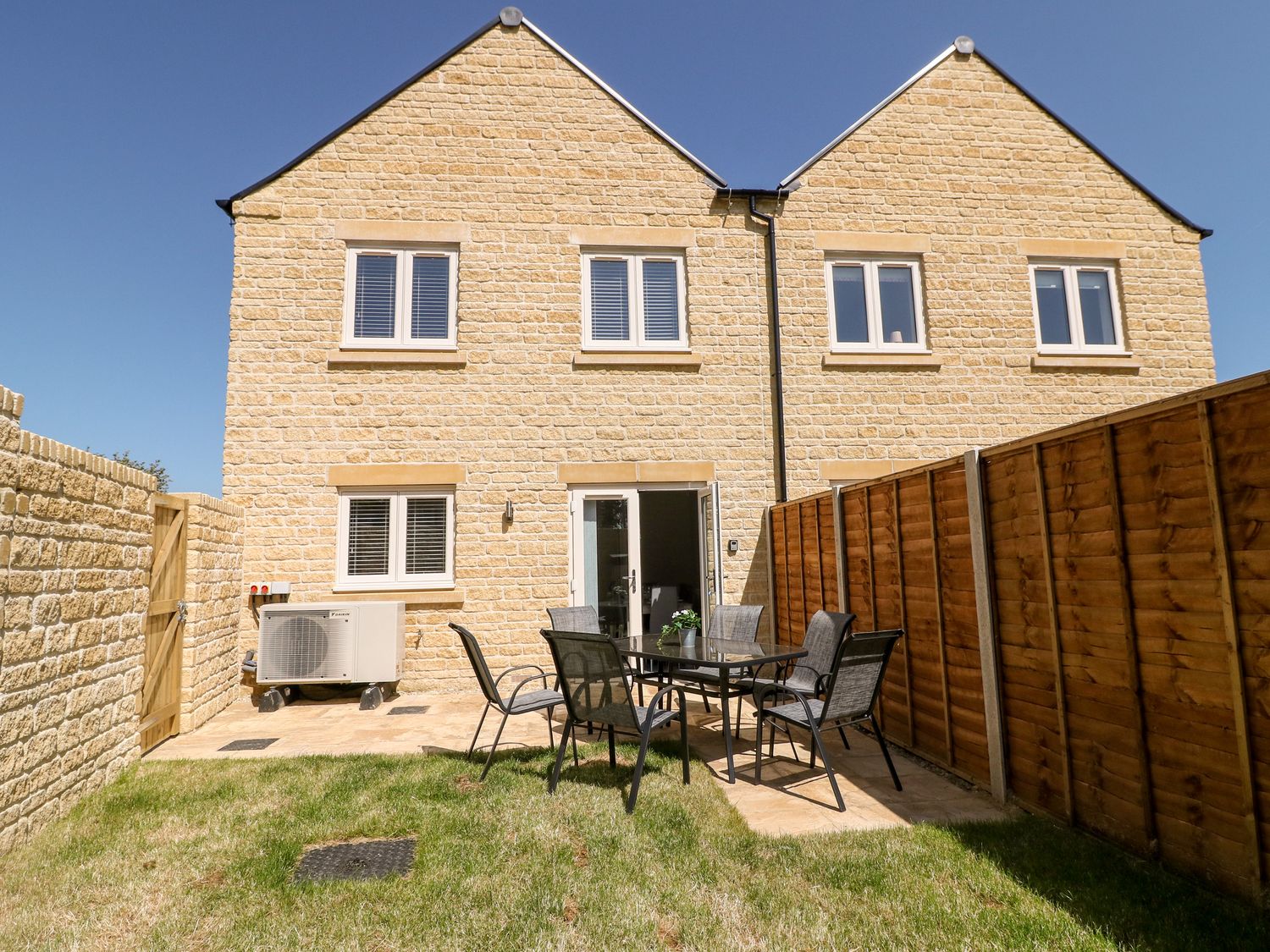 A backyard with a glass table and six chairs on a stone patio behind a brick house at Beau's House in Upper Rissington near Bourton-On-The-Water