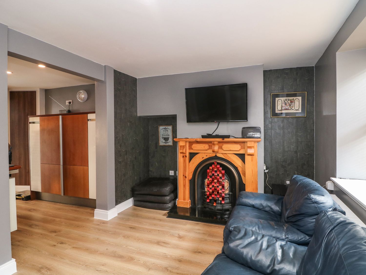 A living room with a blue sofa a wooden fireplace with a television above and an open kitchen area at 78 King Street in Waberthwaite