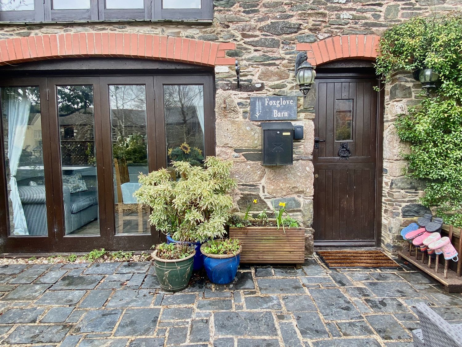 The exterior of a stone building with a dark wooden door and large glass windows with plants in pots and a doormat at Foxglove Barn in South Brent