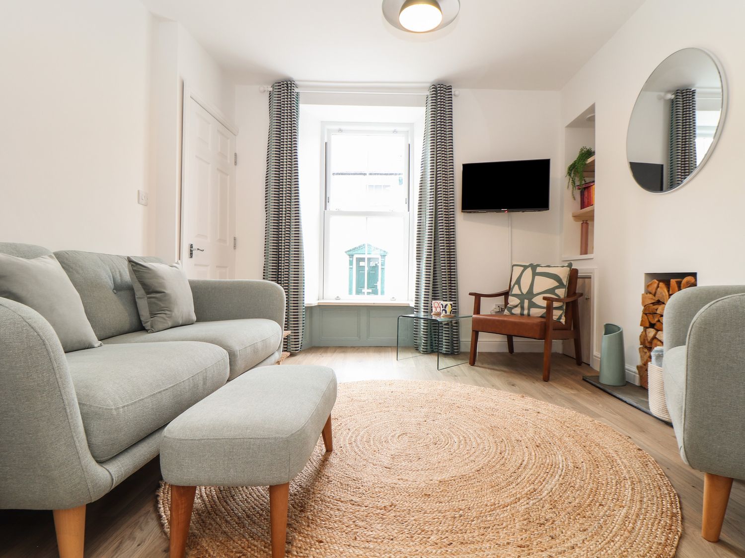 A living room with two grey sofas a round woven rug a wooden chair a glass table a wall mounted tv and a window with curtains at The Lookout in Penzance
