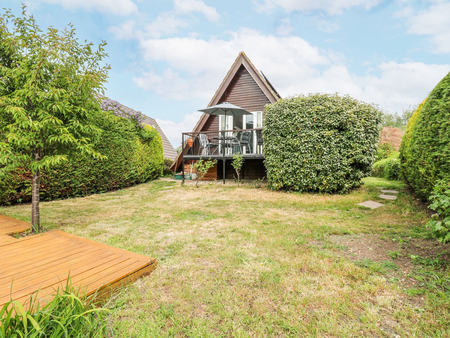 A garden with grass lawn and bushes with a wooden deck and an a frame house with a balcony and outdoor furniture at Kingfisher in Isleham