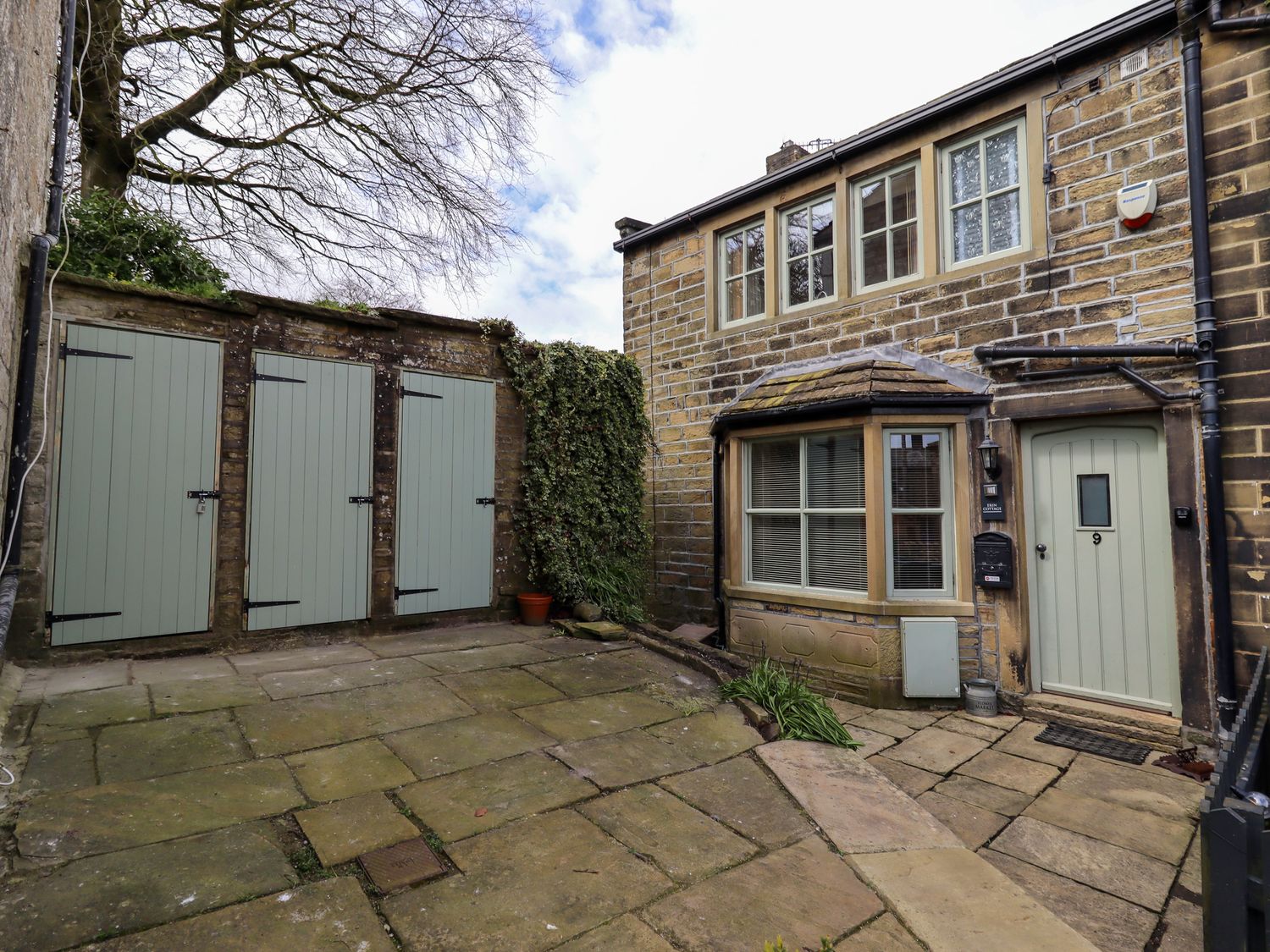 Stone paved courtyard with three green wooden doors and a stone house with bay window and green front door at Erin Cottage in Haworth