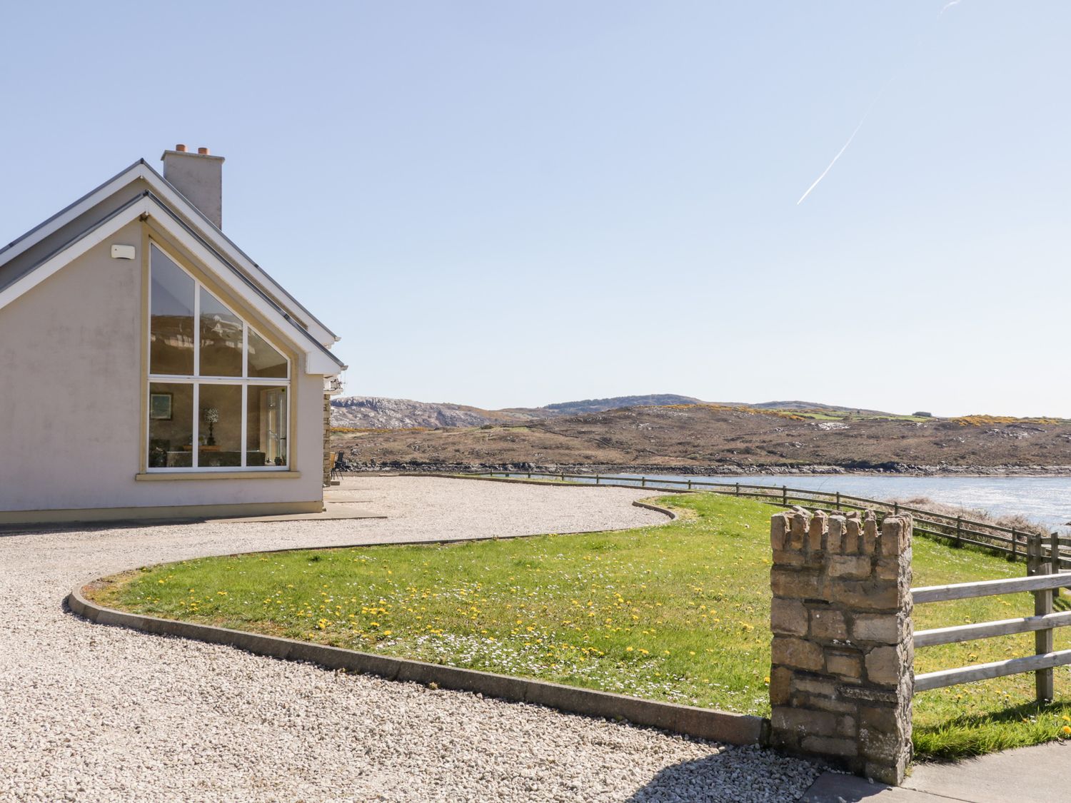 A house with large windows and gravel driveway next to a grassy area by a lake at Inverbeg Cottage 2 in Dundooan Lower near Downings
