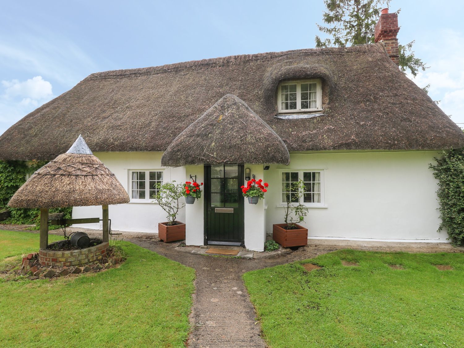 A thatched roof cottage with a front door, two windows, flower pots, a brick well, and a grass lawn at Thatch Cottage in Buckland Newton