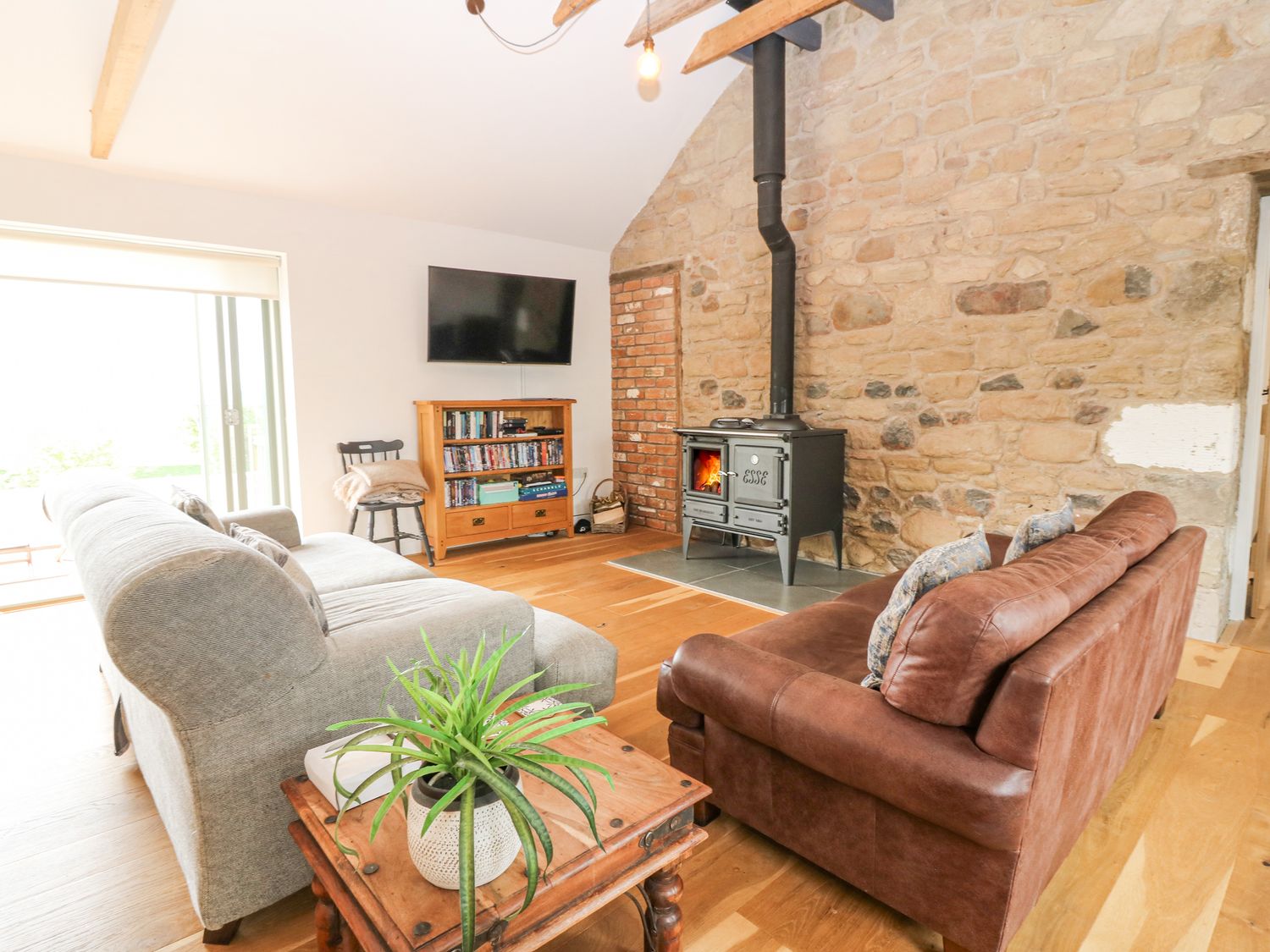 A living room with a gray sofa a brown sofa a wood stove a wooden cabinet with books and a television at Cartshed Ednam near Kelso