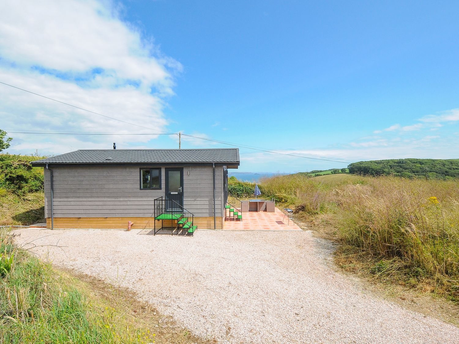 A small gray house with green steps and a tiled patio in a rural area with hills and blue sky at Midway Lodge in Cawsand near Kingsand
