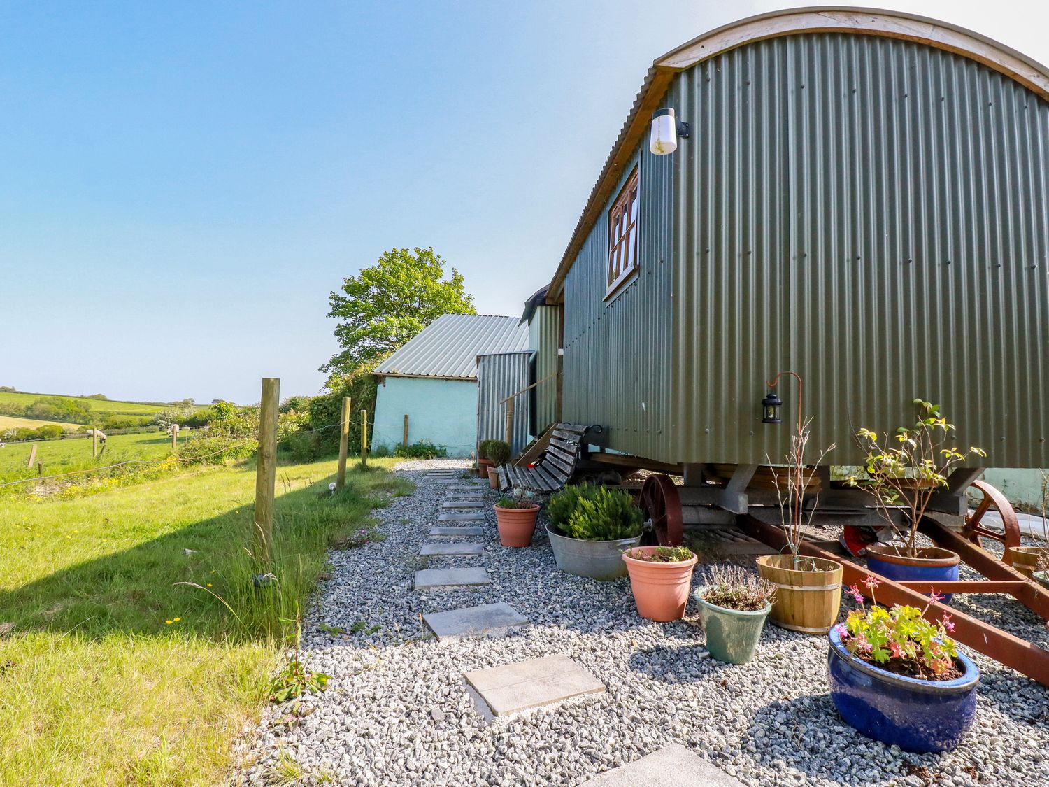 An outdoor space with a corrugated metal hut on wheels a gravel path with potted plants and a bench in a rural area at Puddle Duck Shepherds Hut in Crackington Haven