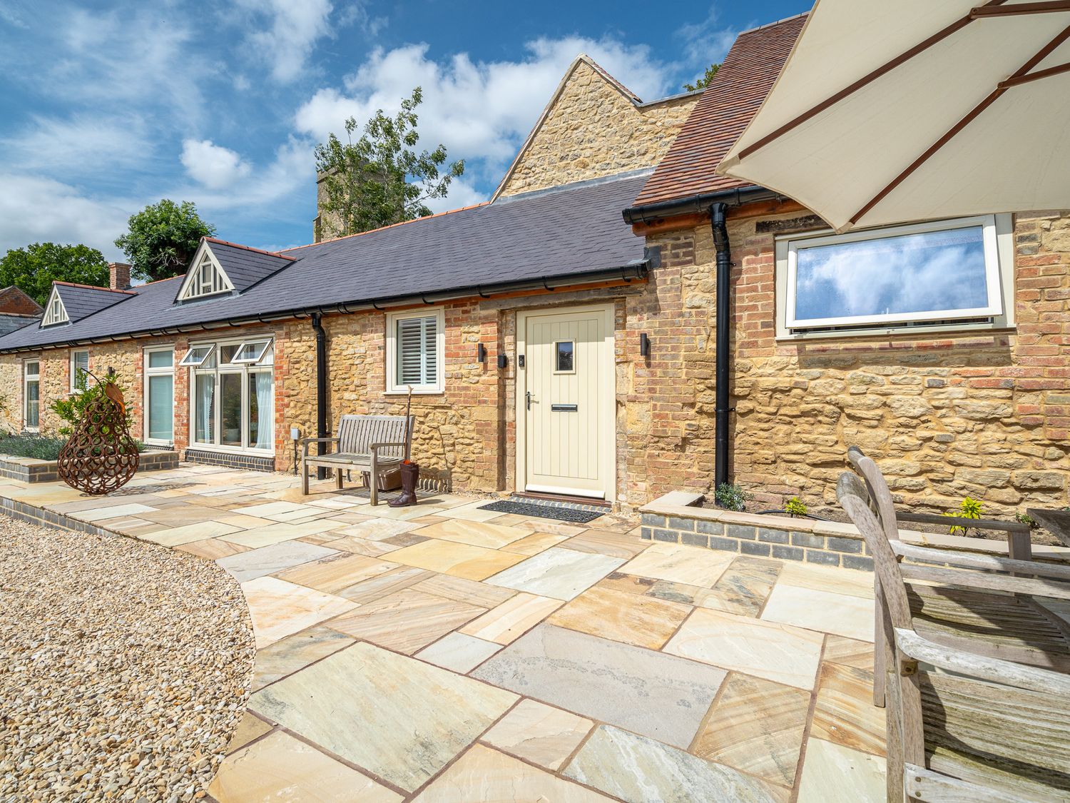 Stone exterior of a house with windows a door benches a large patio umbrella and paved patio at The Stables in Wheatley