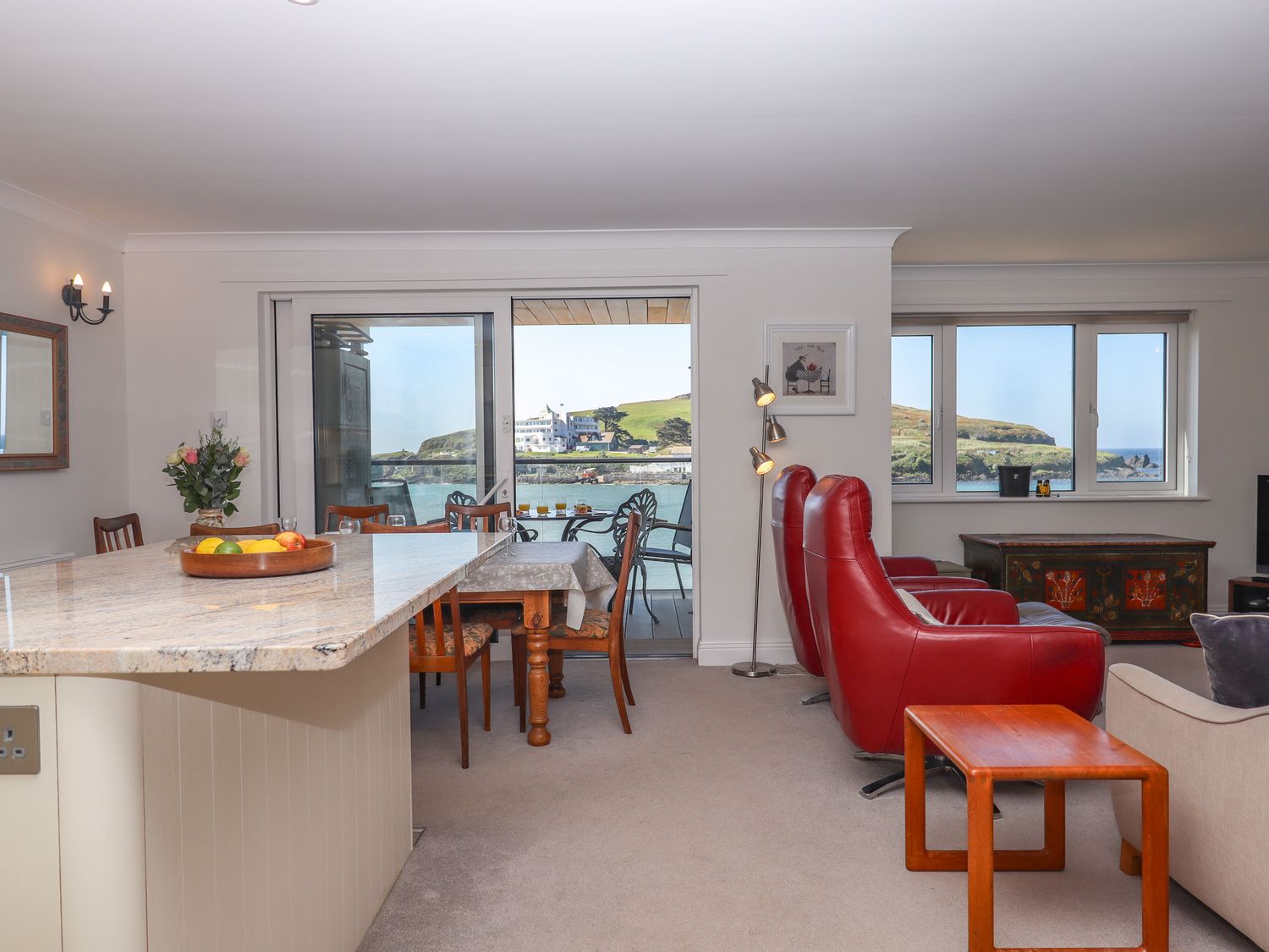 A living and dining area with red leather chairs a wooden table and a granite countertop with a sea view at 14 Burgh Island Causeway in Bigbury-On-Sea