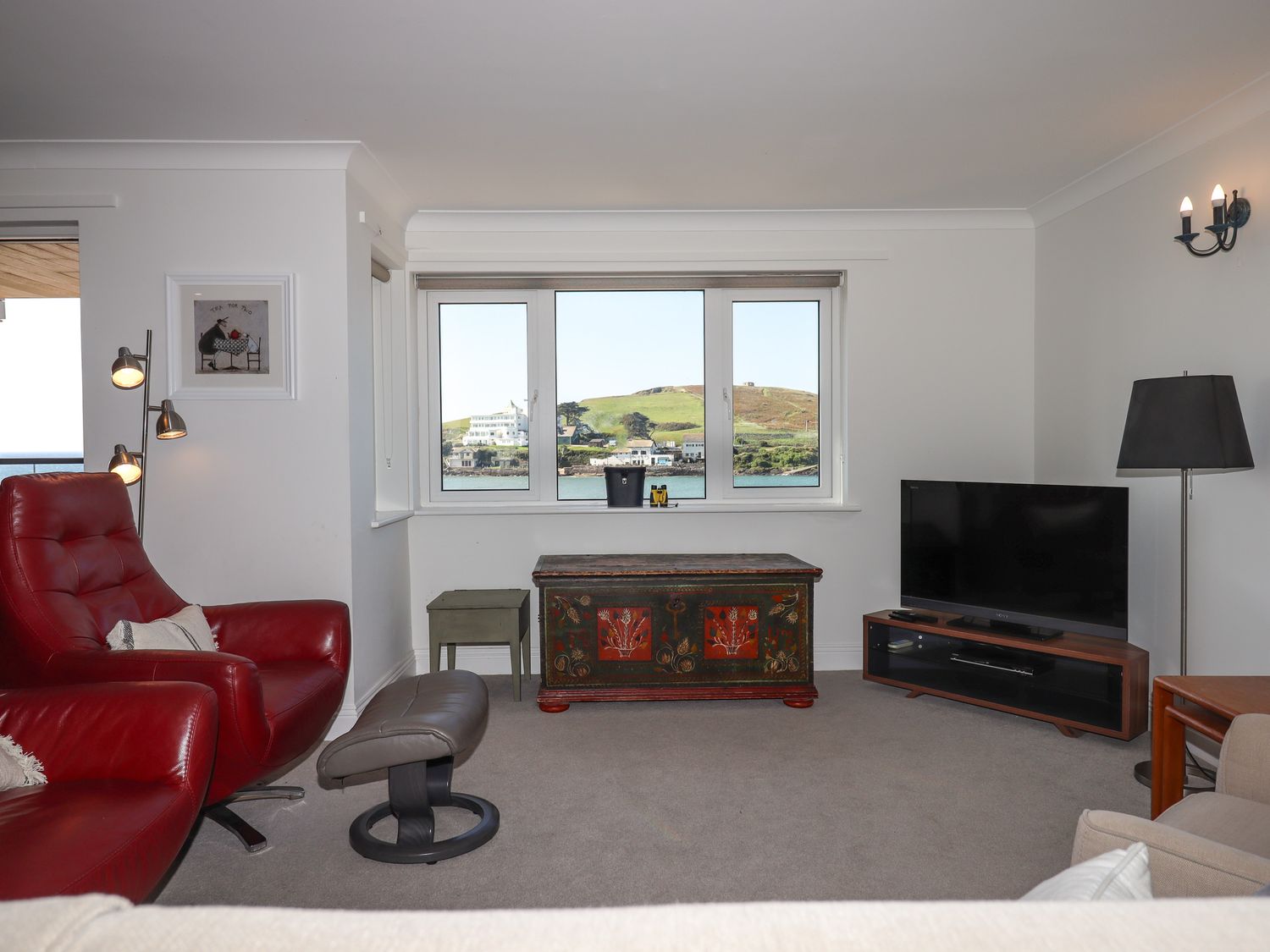 A living room with a red armchair gray footstool antique chest and television at 14 Burgh Island Causeway in Bigbury-On-Sea