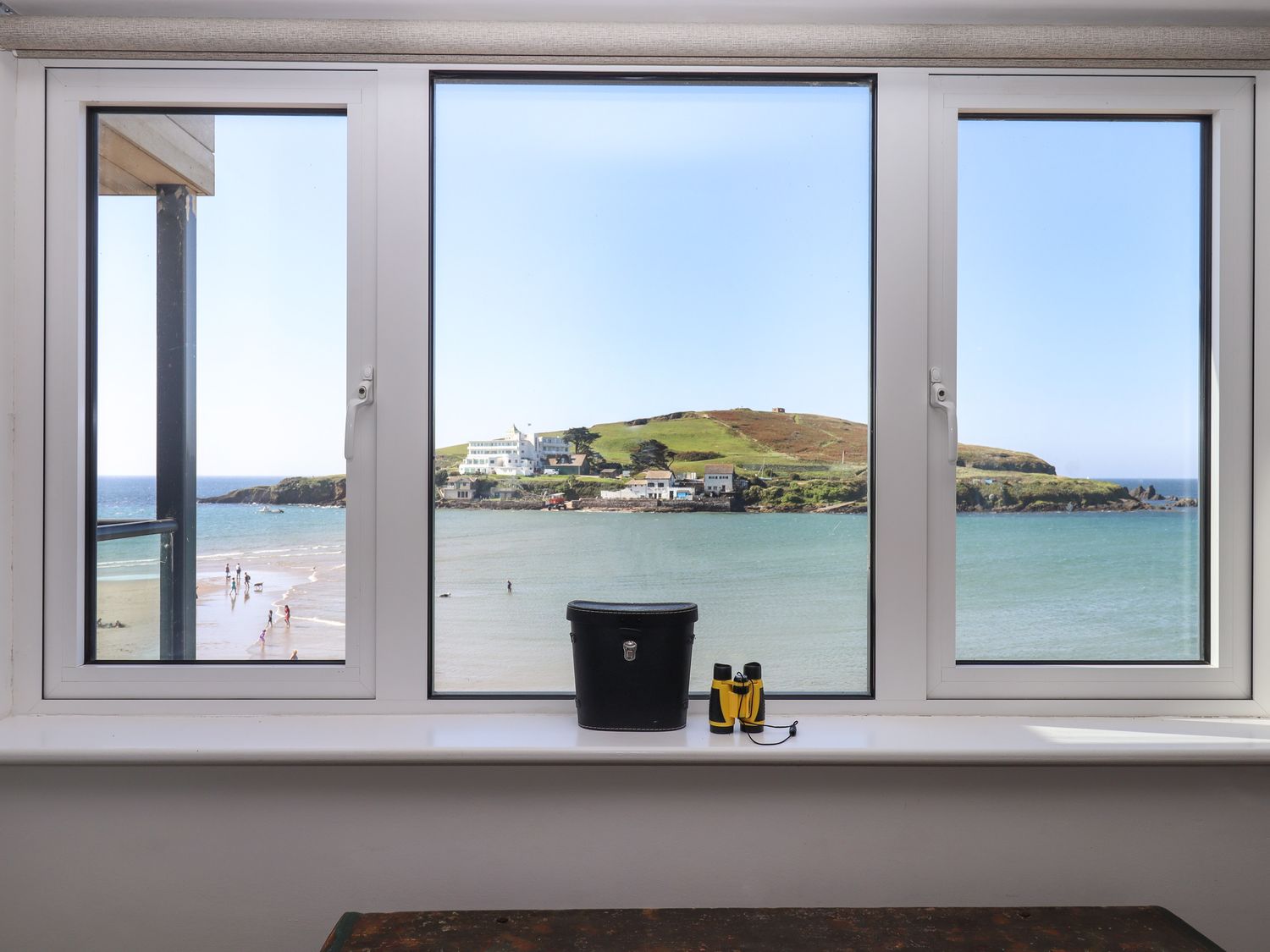 A window with a view of the sea and an island with buildings and grassy hills at 14 Burgh Island Causeway in Bigbury-On-Sea
