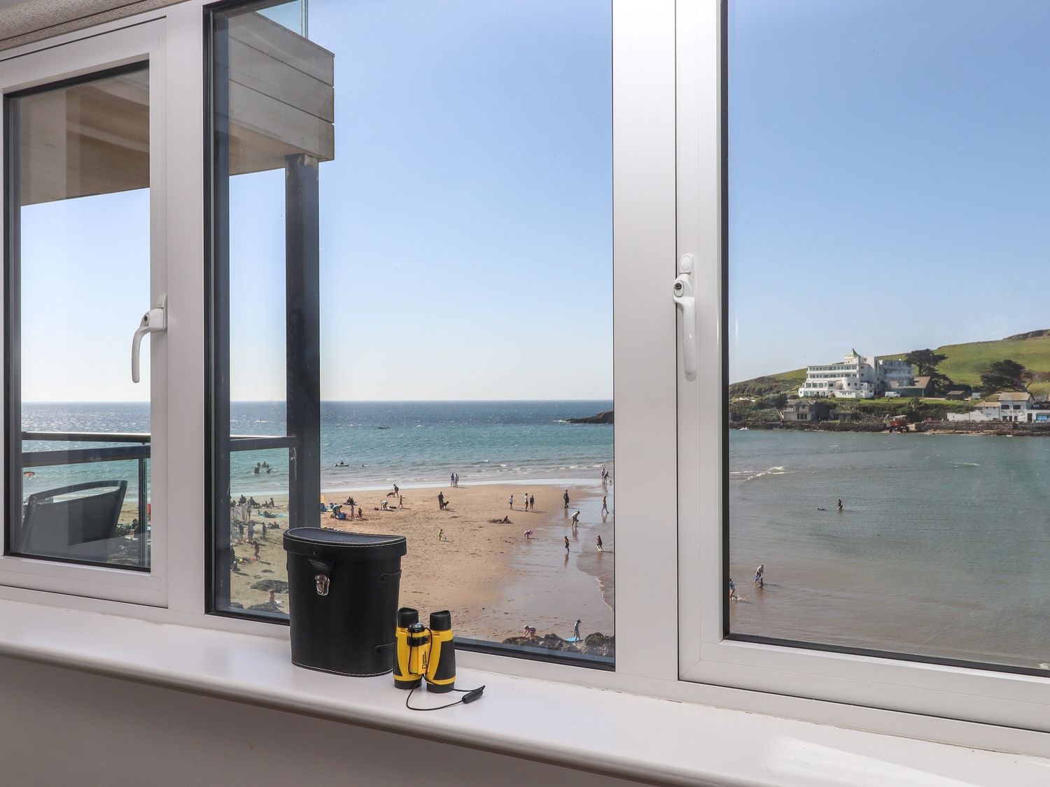 A window view of a beach with people on the sand and a body of water with buildings on a hill at 14 Burgh Island Causeway in Bigbury-On-Sea