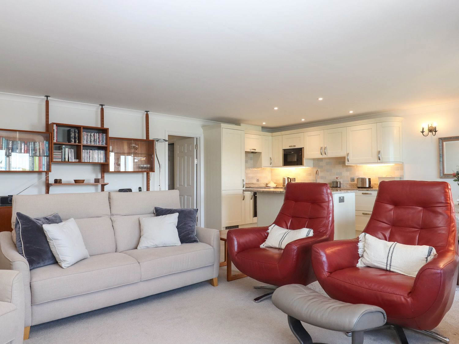 A living room with a beige sofa and two red armchairs facing a kitchen with white cabinets at 14 Burgh Island Causeway in Bigbury-On-Sea