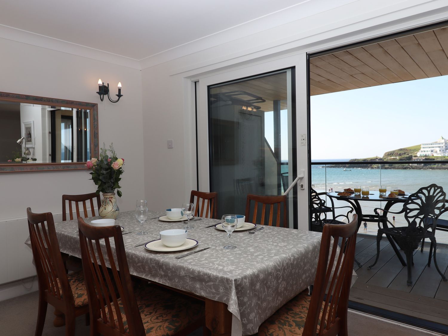 A dining room with a table set for four and a vase of flowers by a sliding glass door opening to a balcony with a view of the sea at 14 Burgh Island Causeway in Bigbury-On-Sea