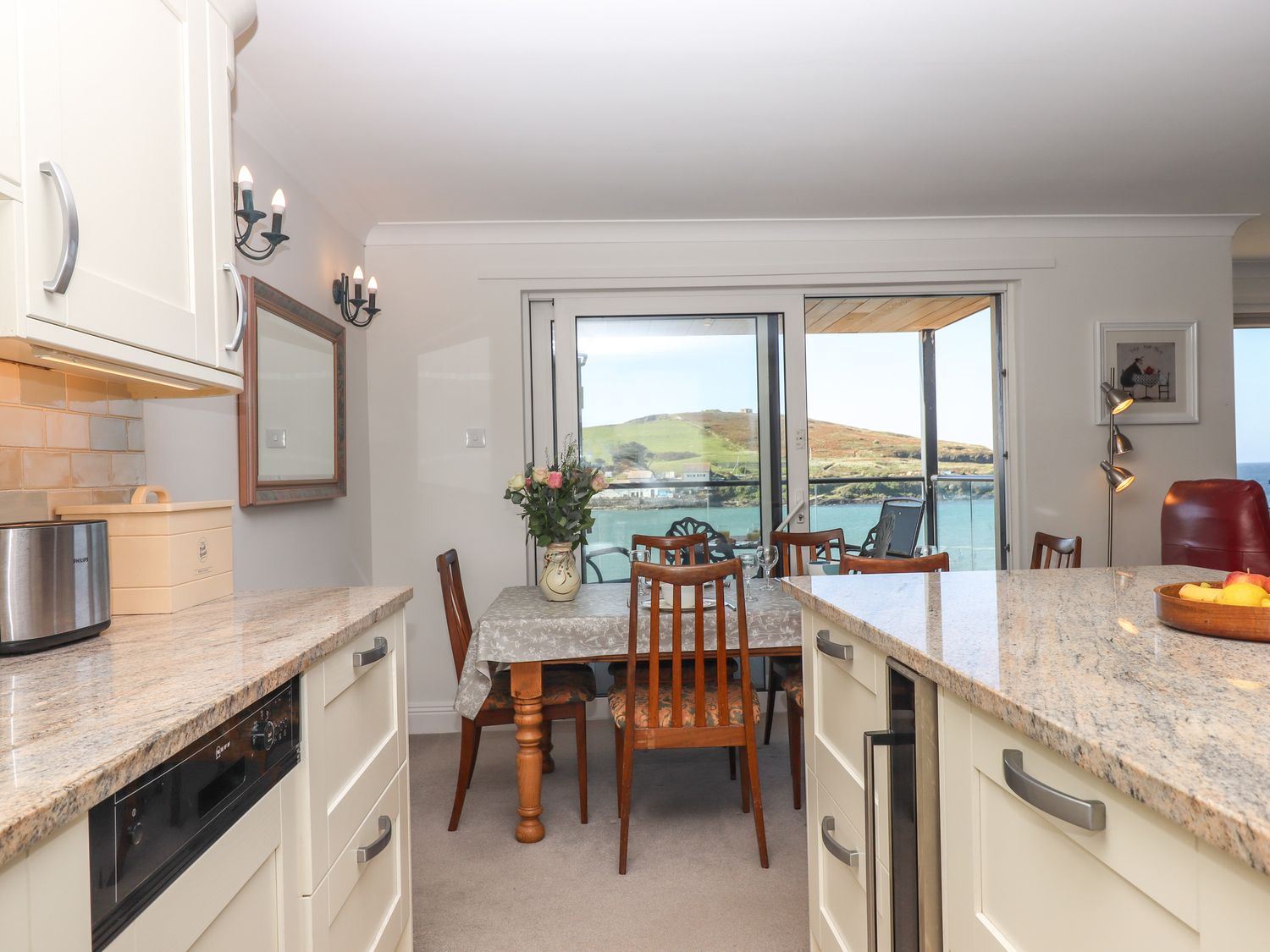 A kitchen and dining area with wooden chairs and a table near a glass door overlooking water and hills at 14 Burgh Island Causeway in Bigbury-On-Sea