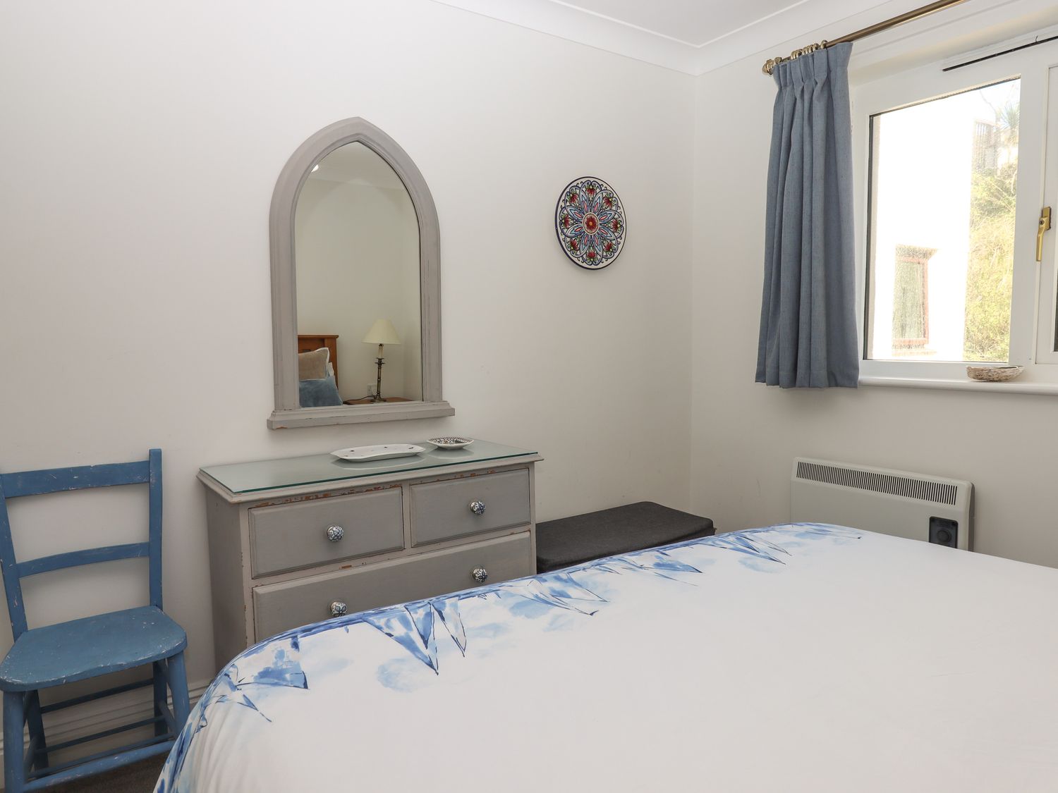 A bedroom with a bed a blue chair a dresser with a mirror and a window with blue curtains at 14 Burgh Island Causeway in Bigbury-On-Sea