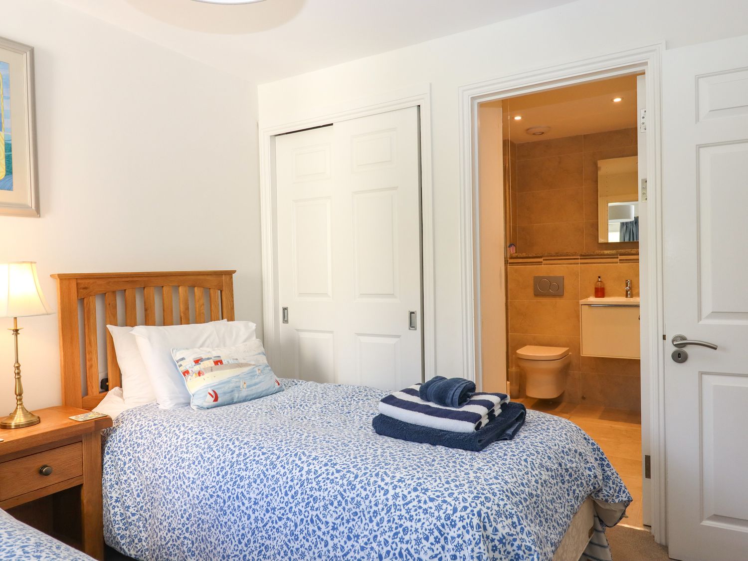 A bedroom with a wooden bed, blue patterned bedding, a bedside table with a lamp, sliding closet doors, and a bathroom visible through an open door at 14 Burgh Island Causeway Bigbury-On-Sea