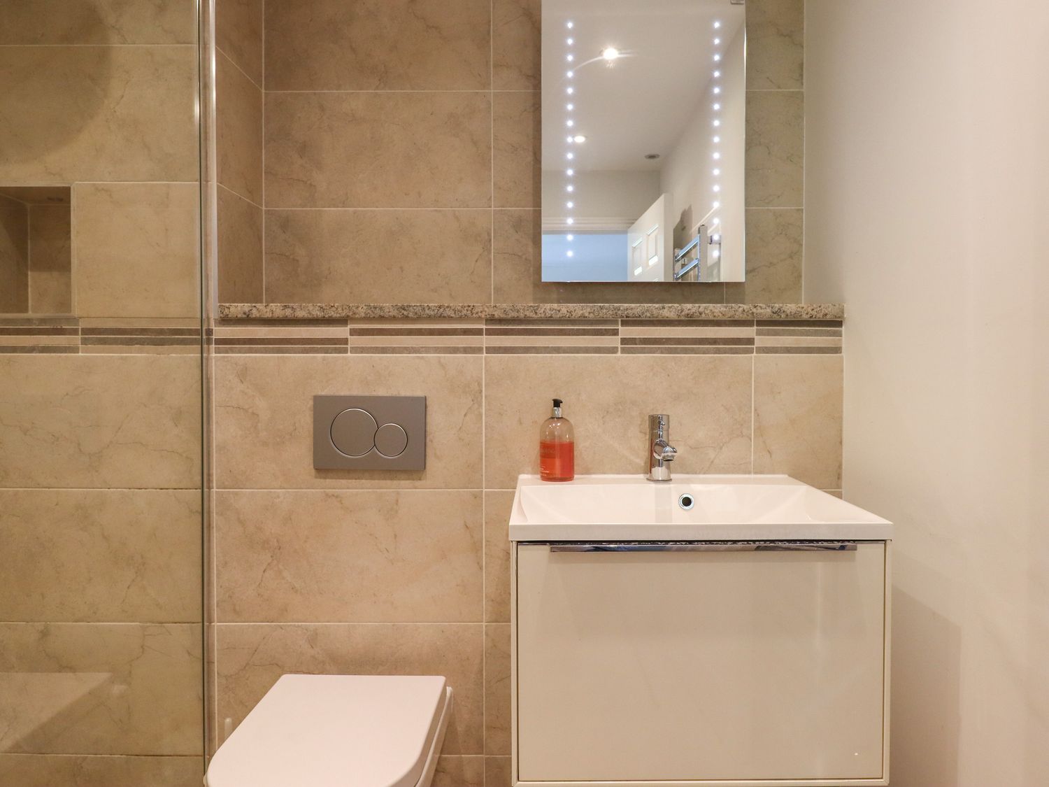 A bathroom with a wall-mounted toilet, a white sink with a cabinet, a mirror, and soap dispenser at 14 Burgh Island Causeway in Bigbury-On-Sea