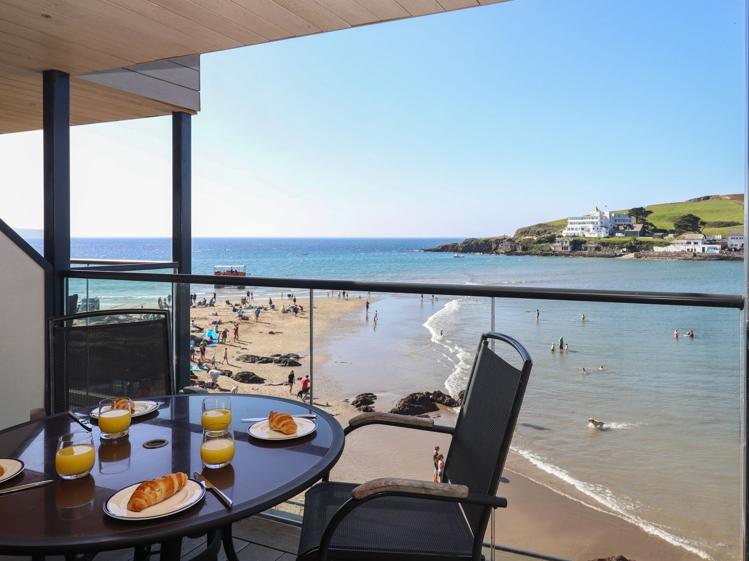 A balcony with a table set with croissants and orange juice overlooking a beach with people swimming and walking at 14 Burgh Island Causeway in Bigbury-On-Sea