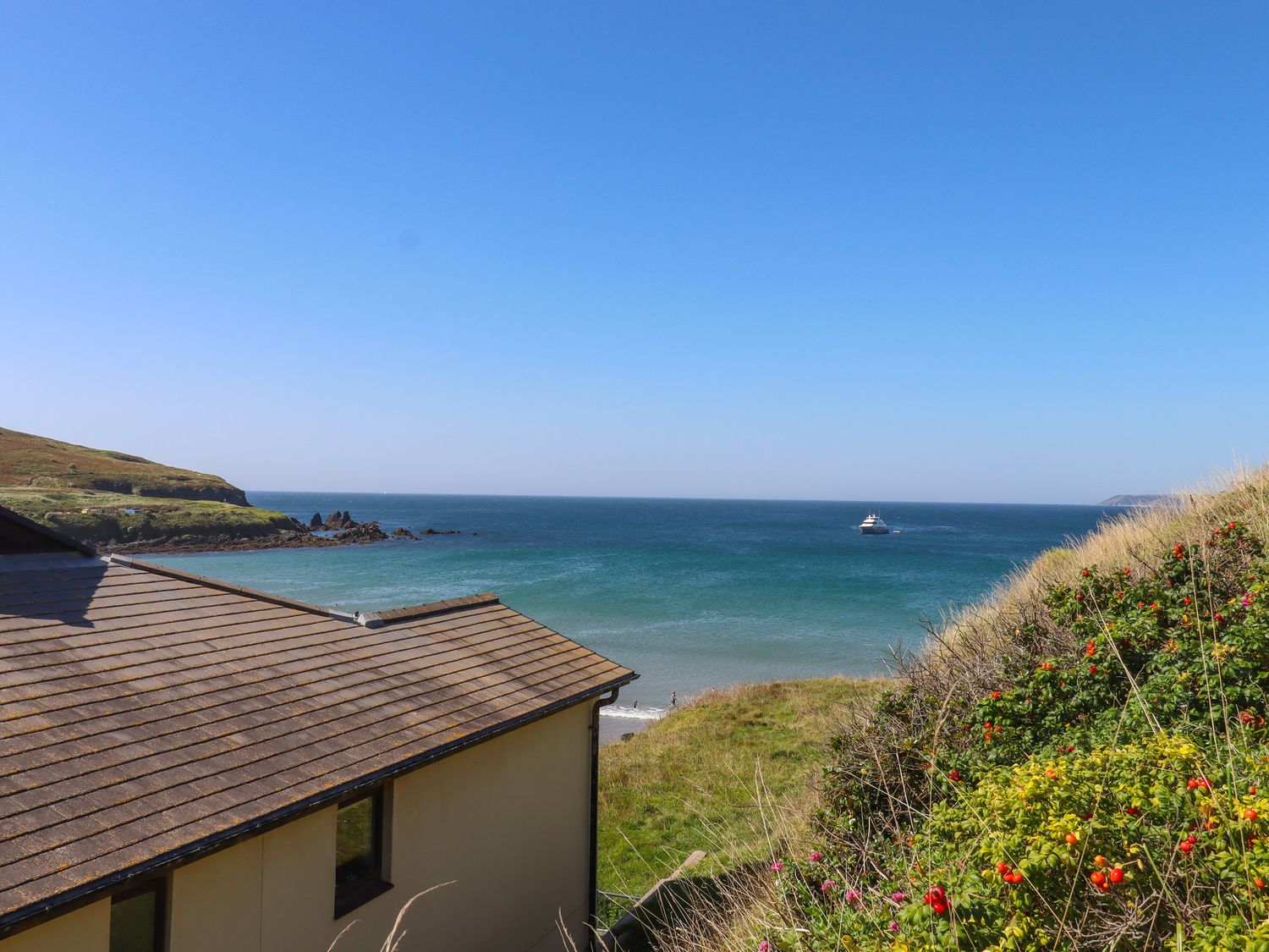 A coastal view with a house roof in the foreground a beach grassy hill with bushes and a boat in the sea at 14 Burgh Island Causeway in Bigbury-On-Sea