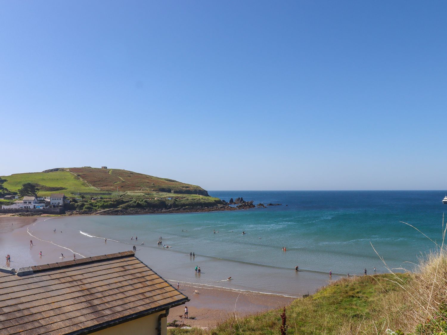 A beach with people walking and swimming near a green hill and houses at 14 Burgh Island Causeway in Bigbury-On-Sea