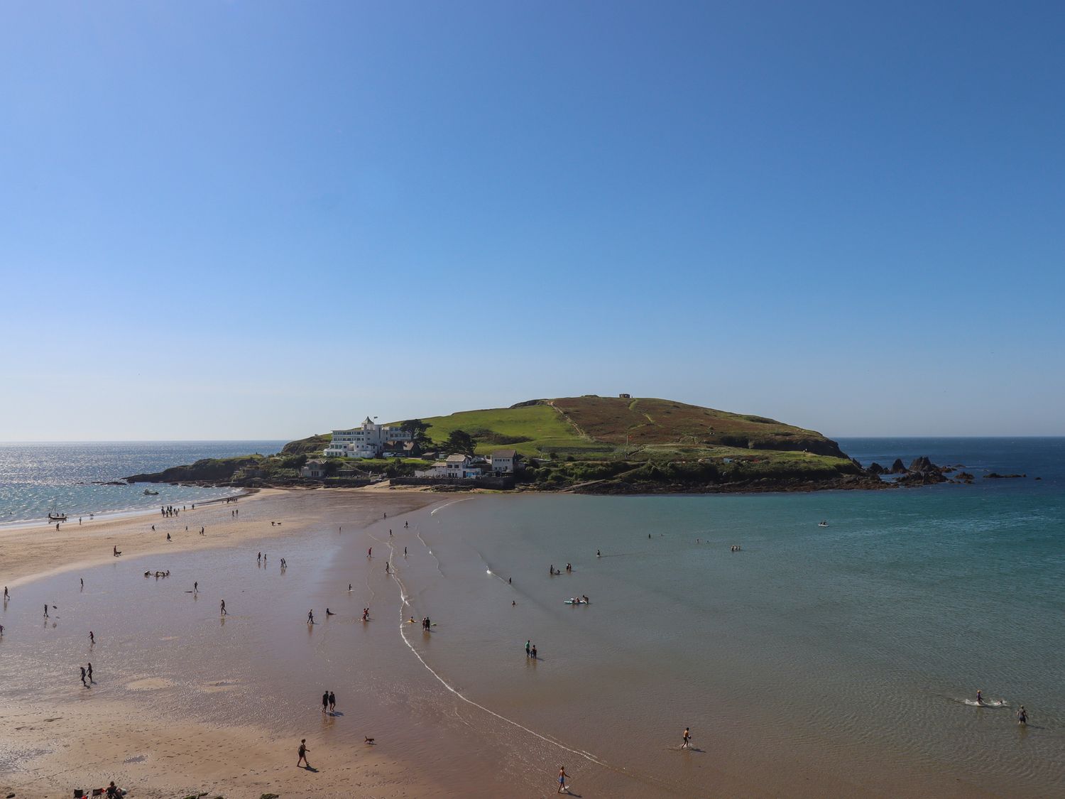 A sandy beach with people walking and swimming with an island and buildings in the background at 14 Burgh Island Causeway in Bigbury-On-Sea