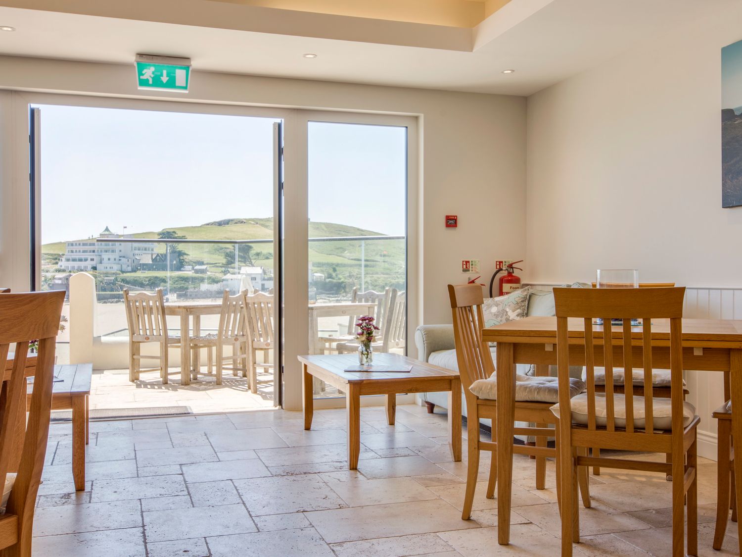 A dining area with wooden chairs and tables near sliding glass doors opening to an outdoor patio with a view of hills and buildings at 14 Burgh Island Causeway in Bigbury-On-Sea