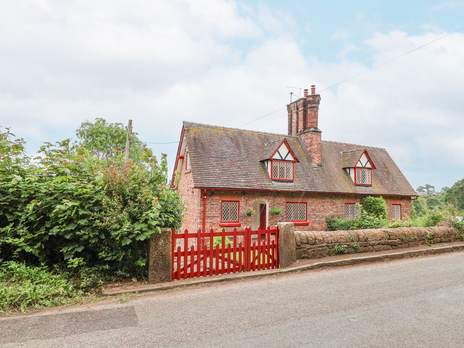 A brick cottage with red window frames and a red gate beside a stone wall at 1 Fountain Cottages in Peckforton Tarporley