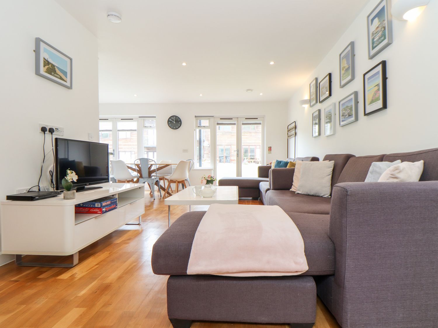 A living room with a brown sectional sofa and cushions a coffee table a tv on a white stand and a dining table with chairs at Garras in Porthtowan