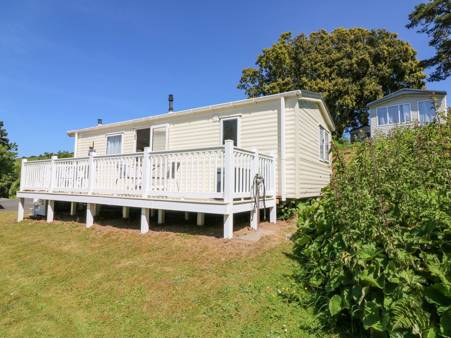 An exterior view of a mobile home with a white deck and railing on a grassy slope with shrubs and trees at Horizon in Combe Martin