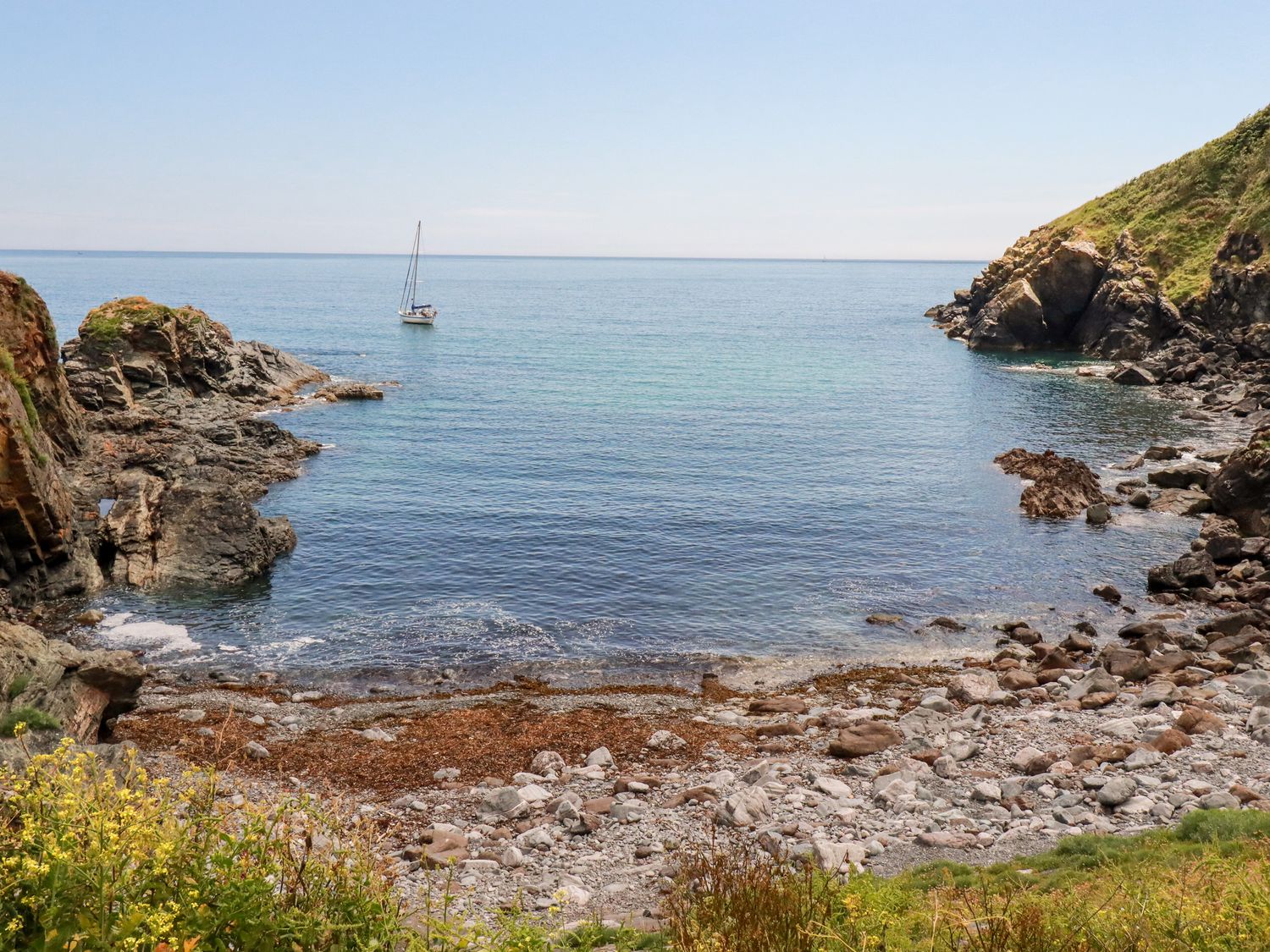 A rocky seaside cove with a sailboat on the water and grassy slopes on either side at Beach Cottage in Cadgwith