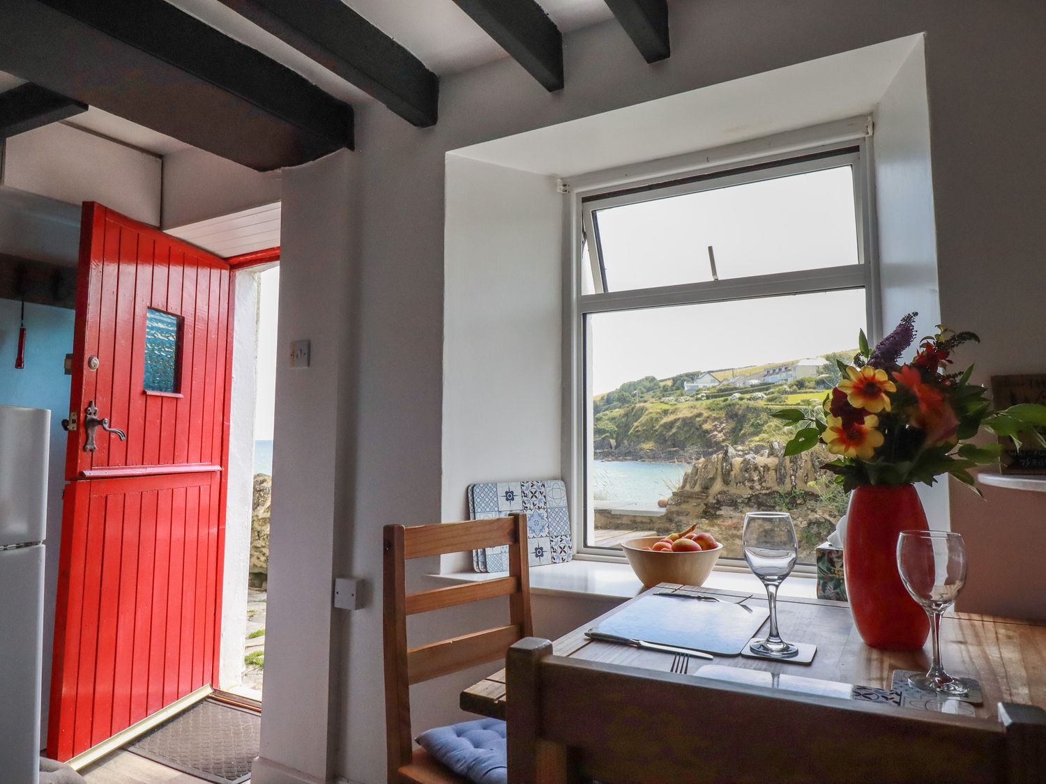 A kitchen dining area with a red door open to the outside and a window with a view of the coastline at Chy Rani in Portmellon near Mevagissey