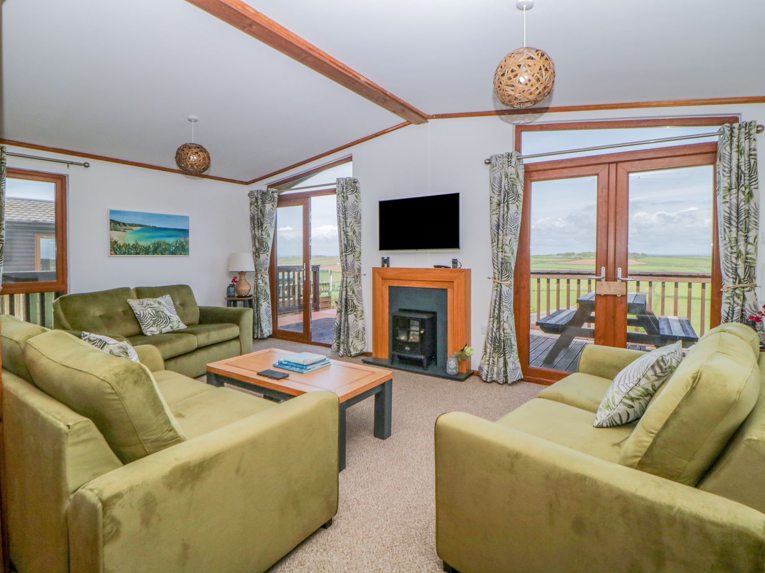 A living room with green sofas a wooden coffee table a wall-mounted TV and glass doors opening to a balcony at Three Views Lodge in Whitsand Bay Fort Holiday Village