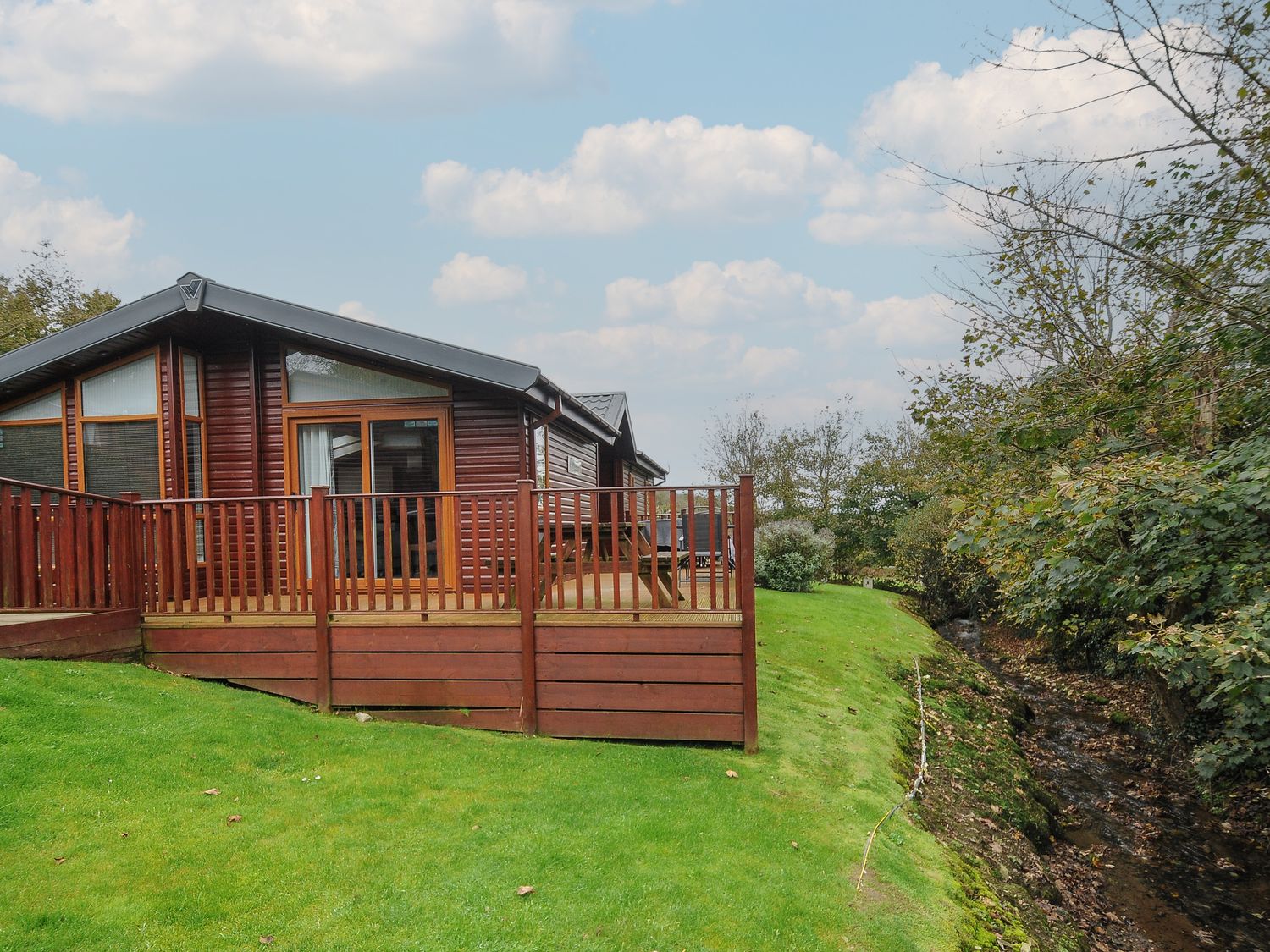 A wooden cabin with a deck and railing on a grassy lawn next to a small creek at Haven Lodge in Bossiney Bay near Tintagel